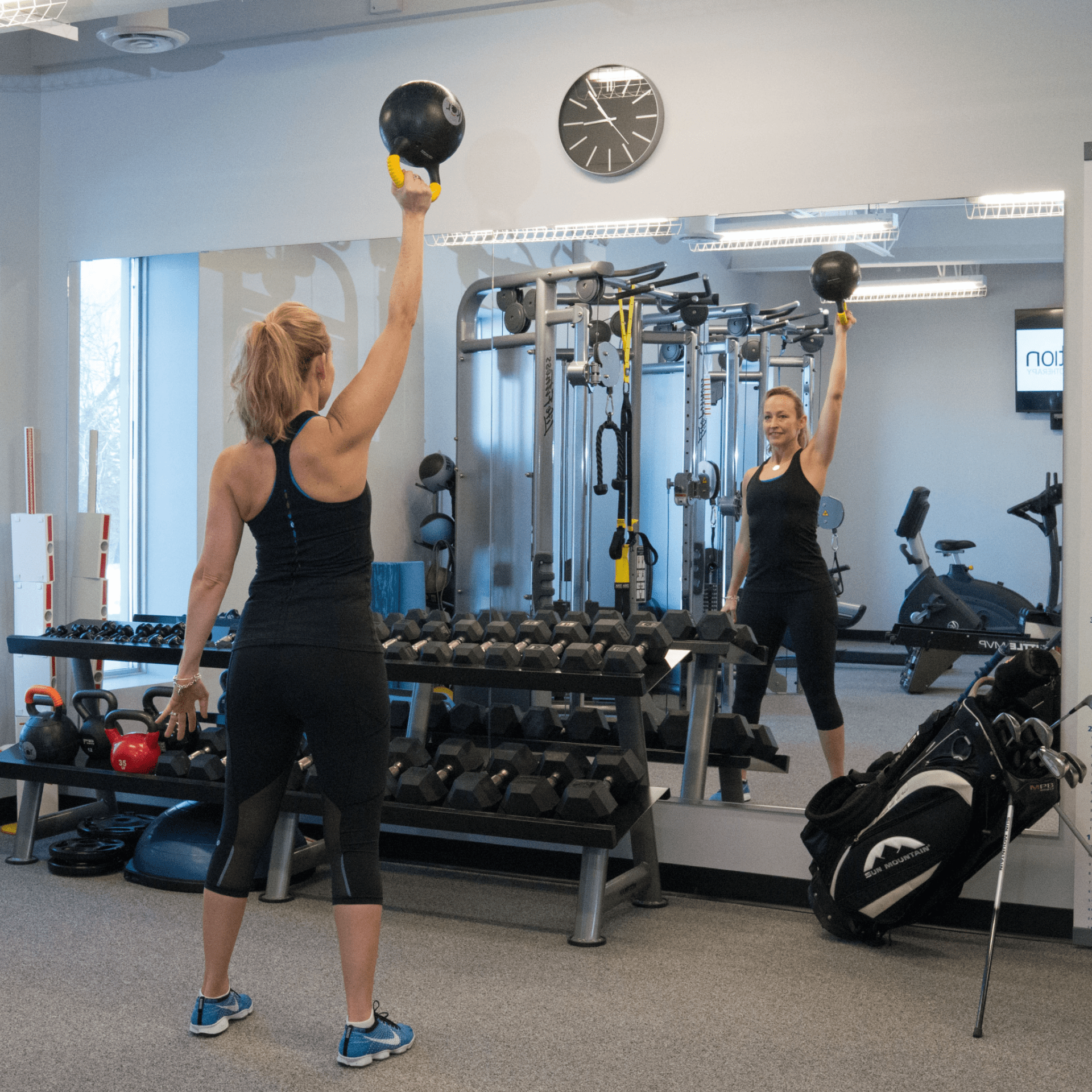 a woman standing in a gym, holding a weight above her head